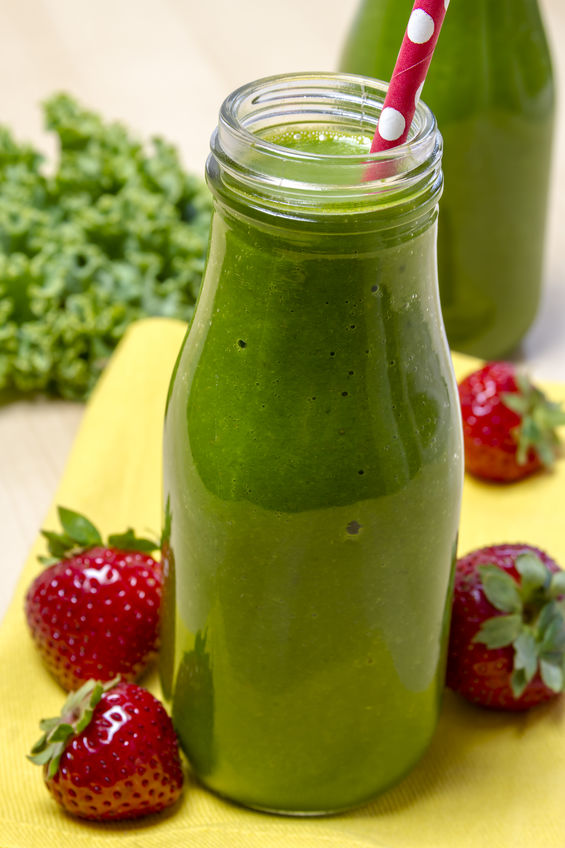 Healthy green juice smoothie in glass bottle sitting on yellow napkin with fresh strawberries and kale