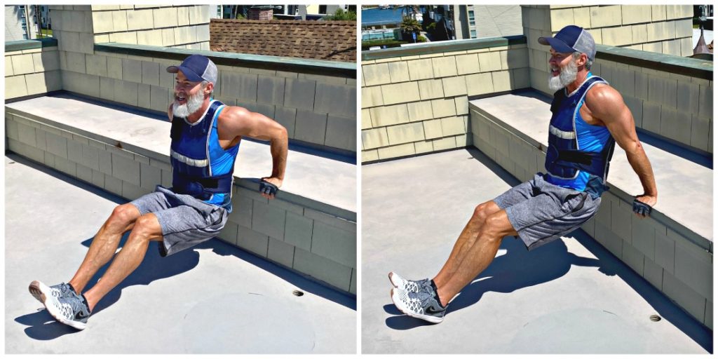 Athlete with silver beard is wearing a weighted vest while doing tricep bench dips.